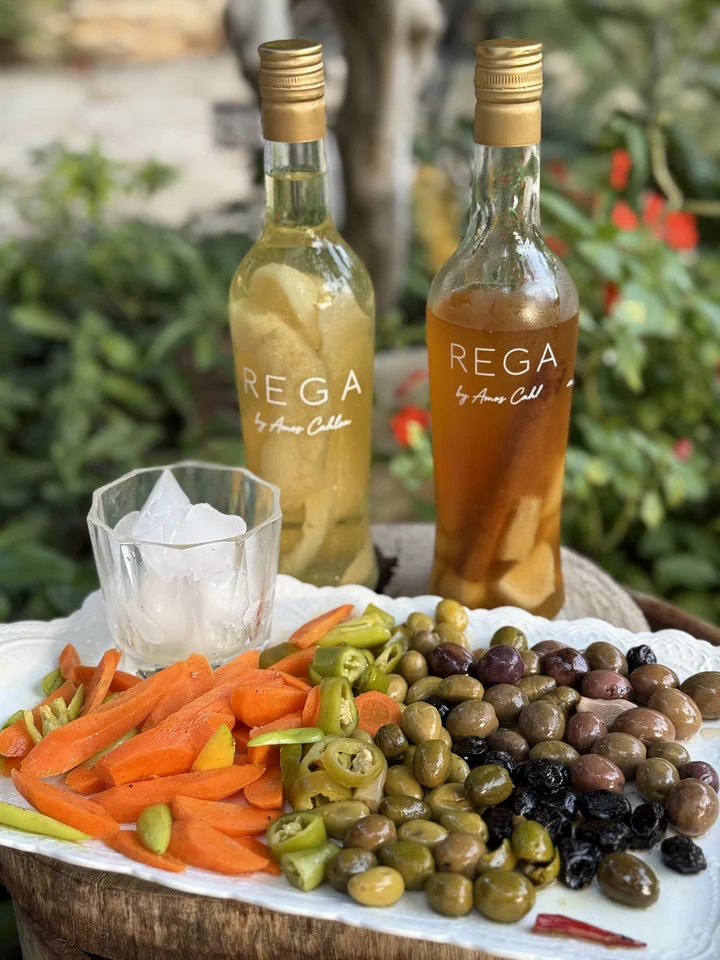 Two chilled REGA bottles in pale yellow and amber, beside an iced glass, olives and vegetables in the garden
