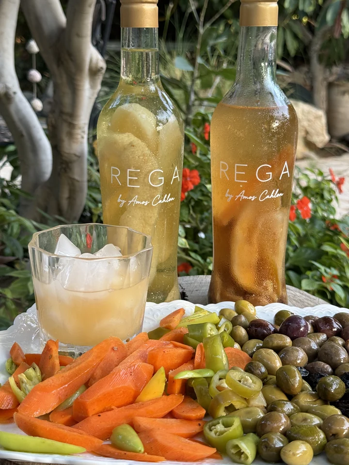 A close view of two REGA bottles with condensation and the Amos Cahlon signature, beside a glass, olives and vegetables