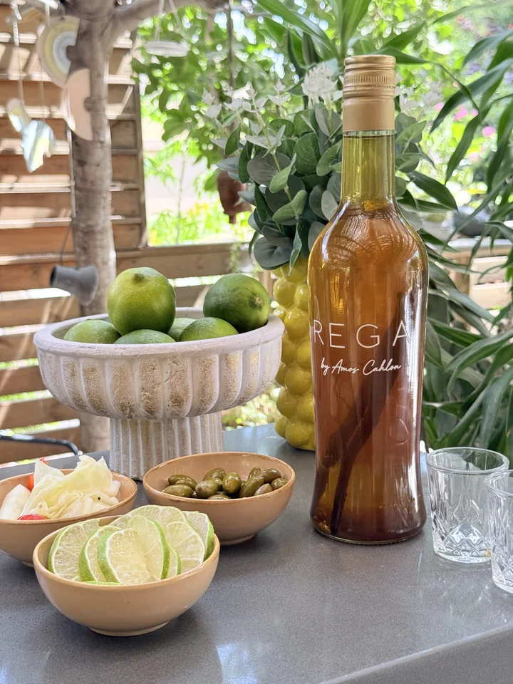 An amber REGA bottle with the REGA name and by Amos Cahlon signature, beside citrus and greenery